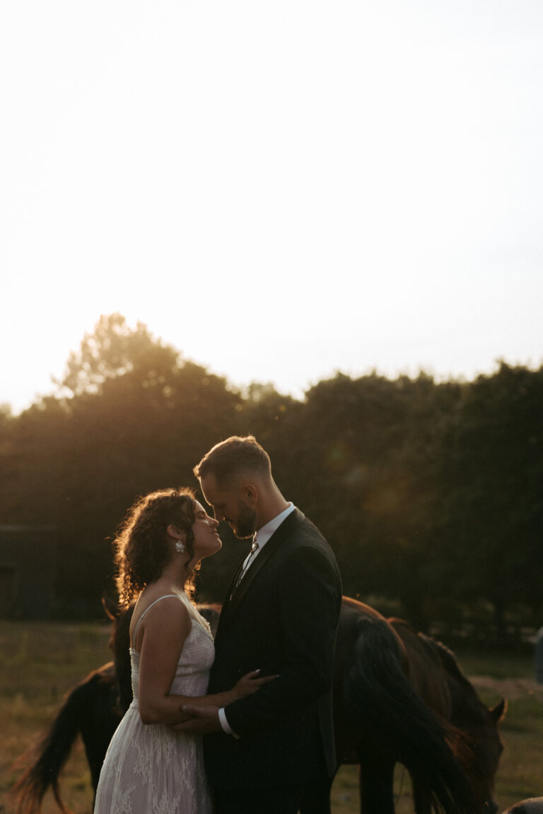Romantic couple portrait in natural light with the bride’s veil flowing and horses grazing in the background.